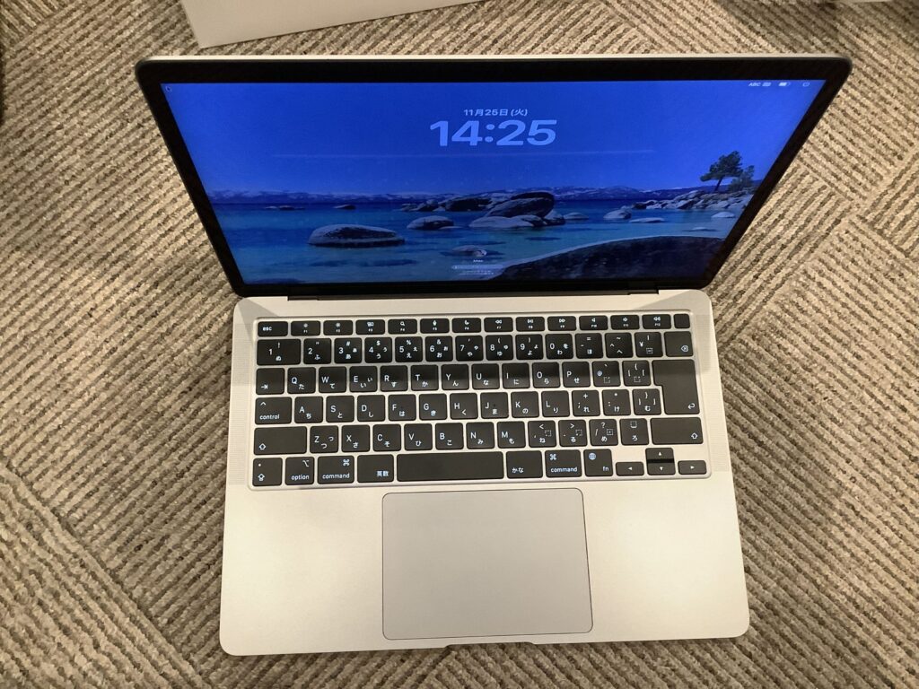 Open silver laptop on a striped carpet, screen shows a blue wallpaper with a large 14:25 time display and a rocky lakeside scene in the background.